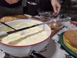Mani che preparano una torta fatta in casa, con una ciotola di crema, una base di pan di Spagna e nocciole tritate.